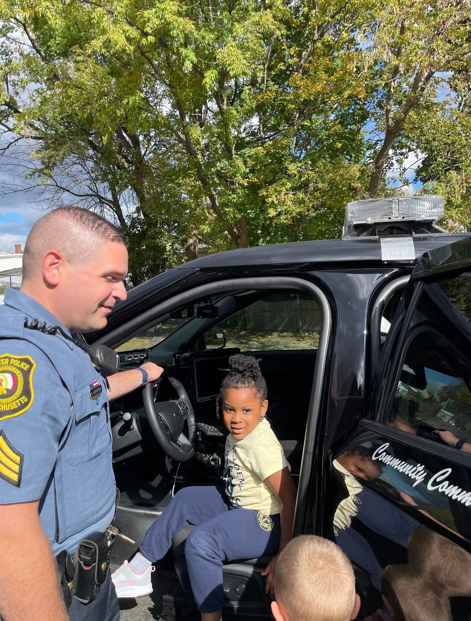 A police officer is standing next to a police car with a little girl sitting in it.