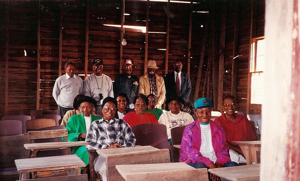 People seated in desks in a wooden room, some standing behind them.