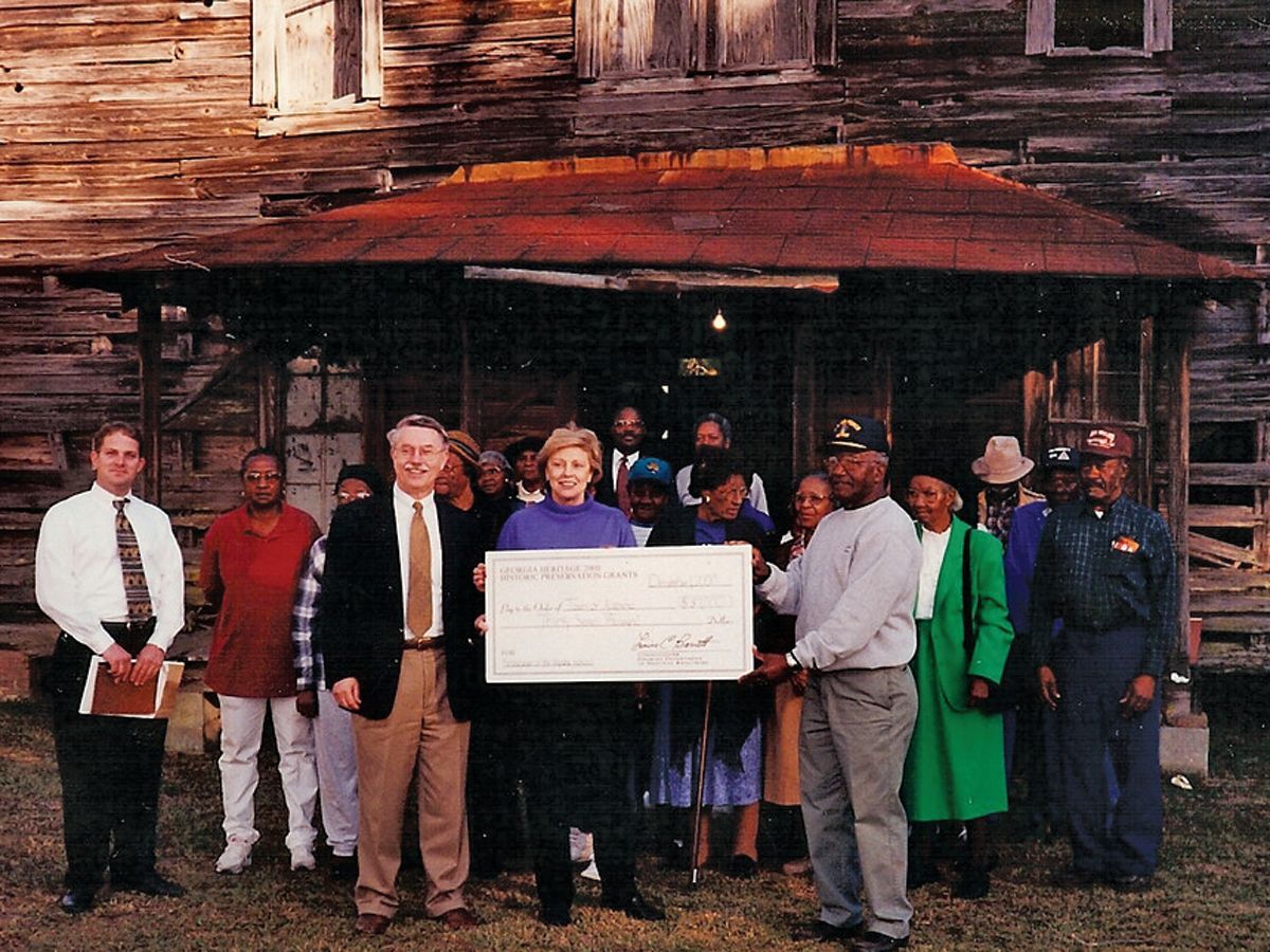 Group of people holding a large check in front of a rustic wooden building with a porch.