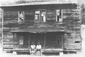 Two men sit on a porch of a weathered, two-story wooden building.