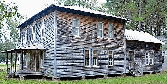 Two-story wooden house with weathered siding and porch; set in grassy area.
