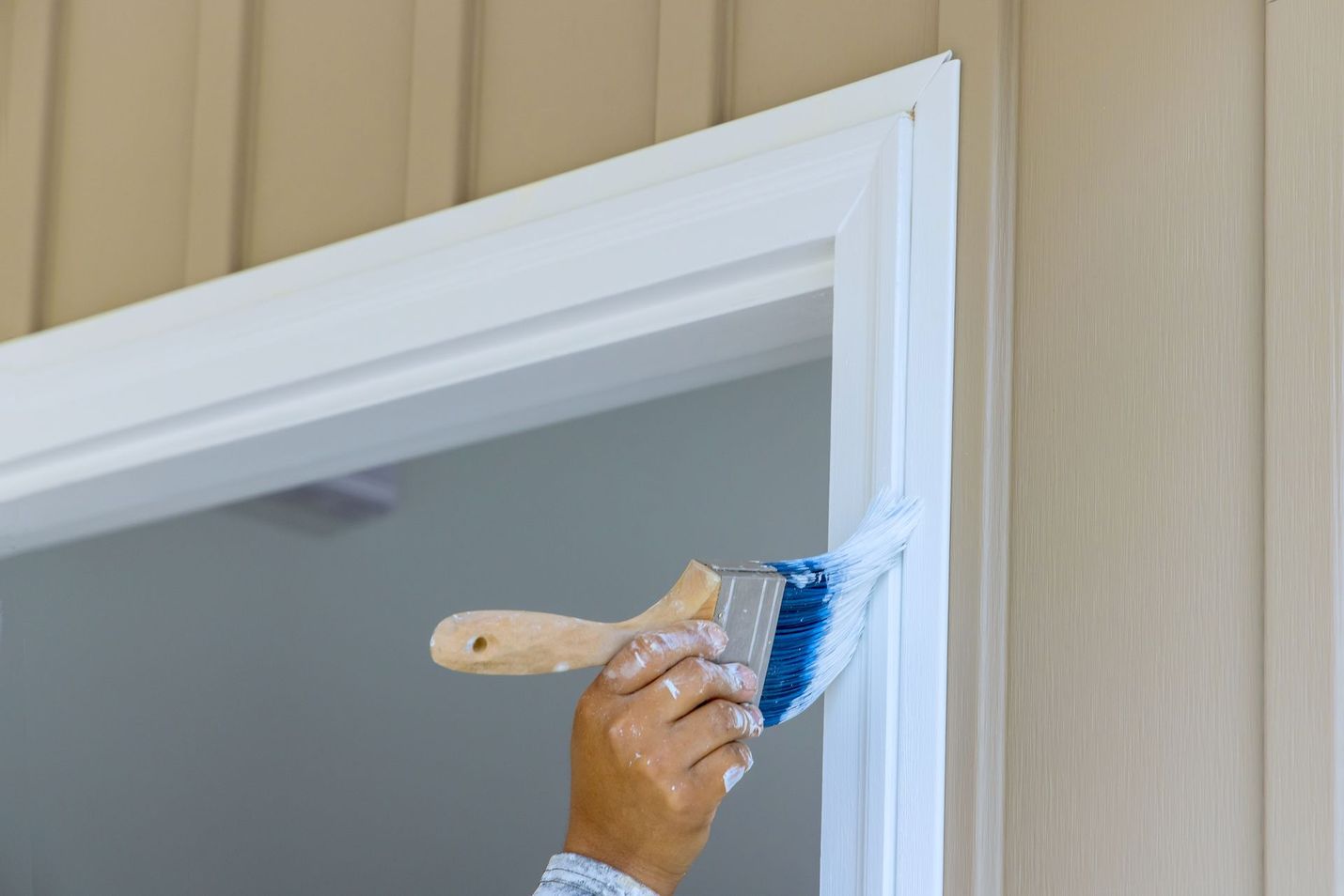 Hand painting a white door frame with a paintbrush. The wall is beige, and the setting appears to be indoors.