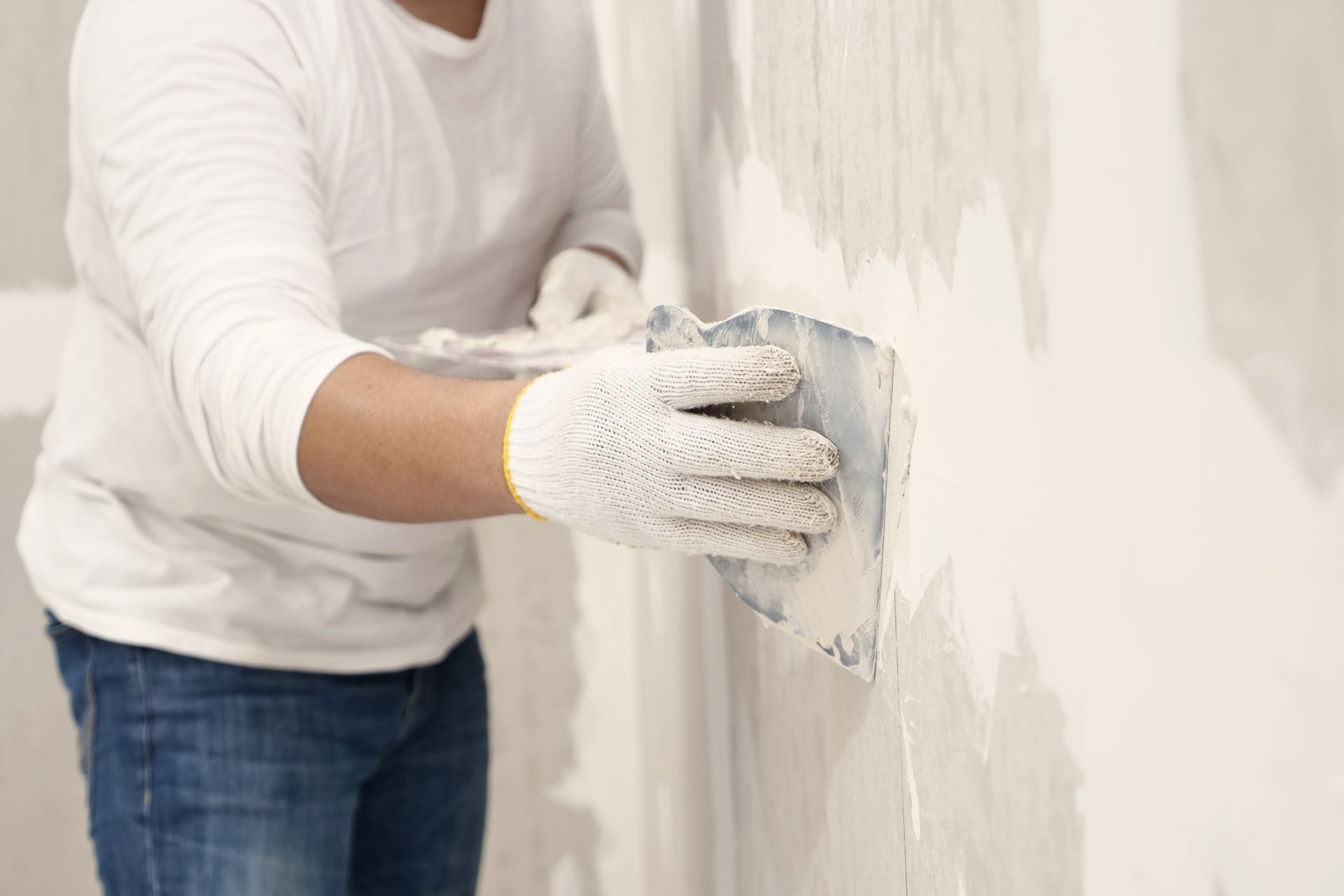 Person wearing gloves and jeans, applying plaster to a white wall with a trowel.