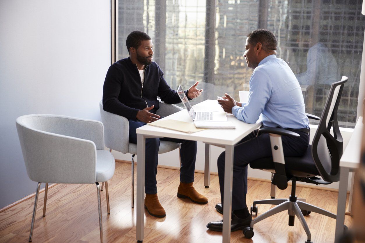 Two people in a bright office meeting at a small round table, talking with papers between them.