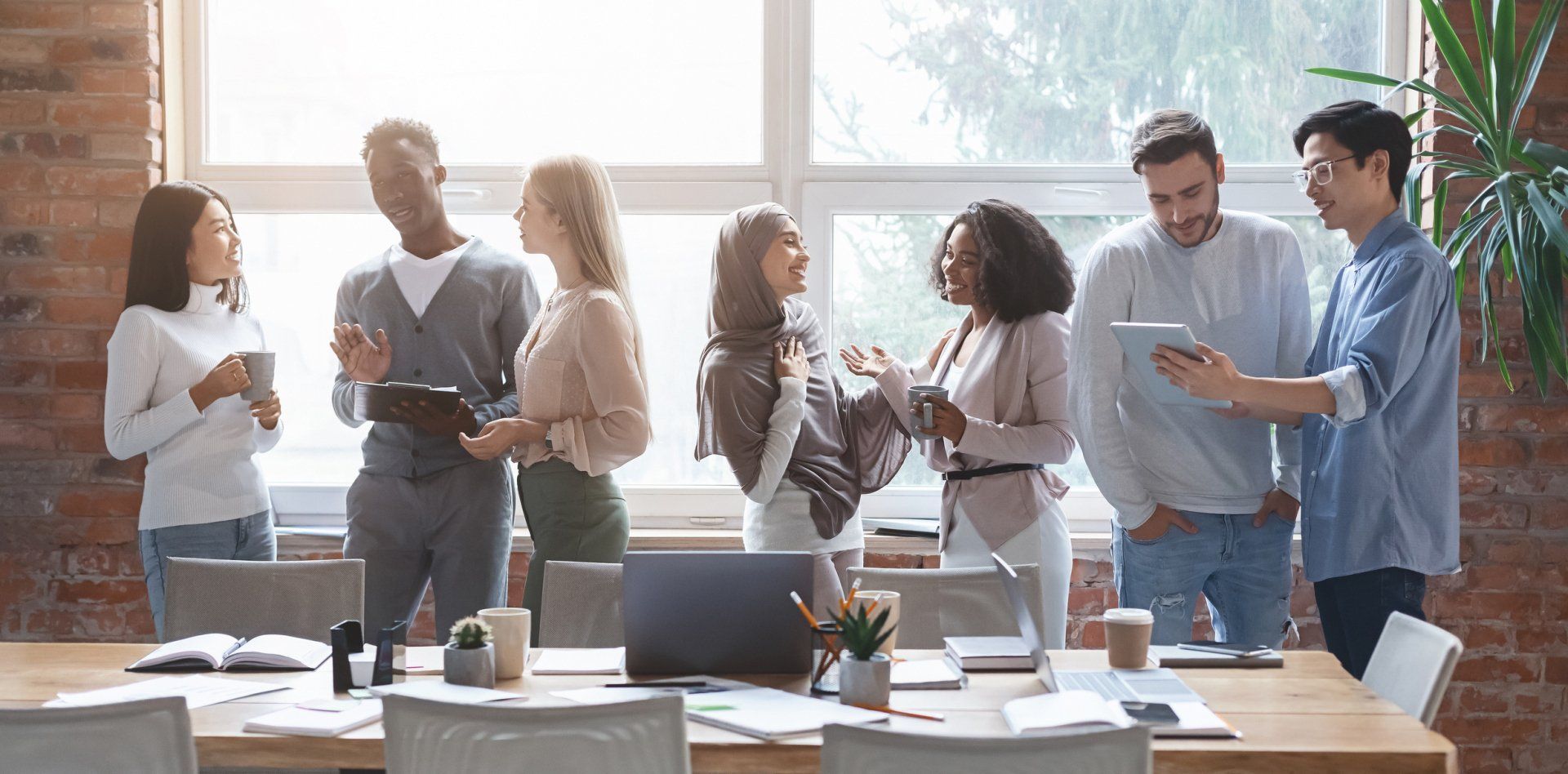 Coworkers chatting and smiling in a bright office meeting, standing around desks with laptops and papers