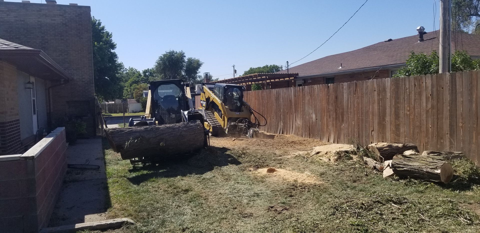 Orange excavator removing a large tree stump from a residential yard. A house is in the background.