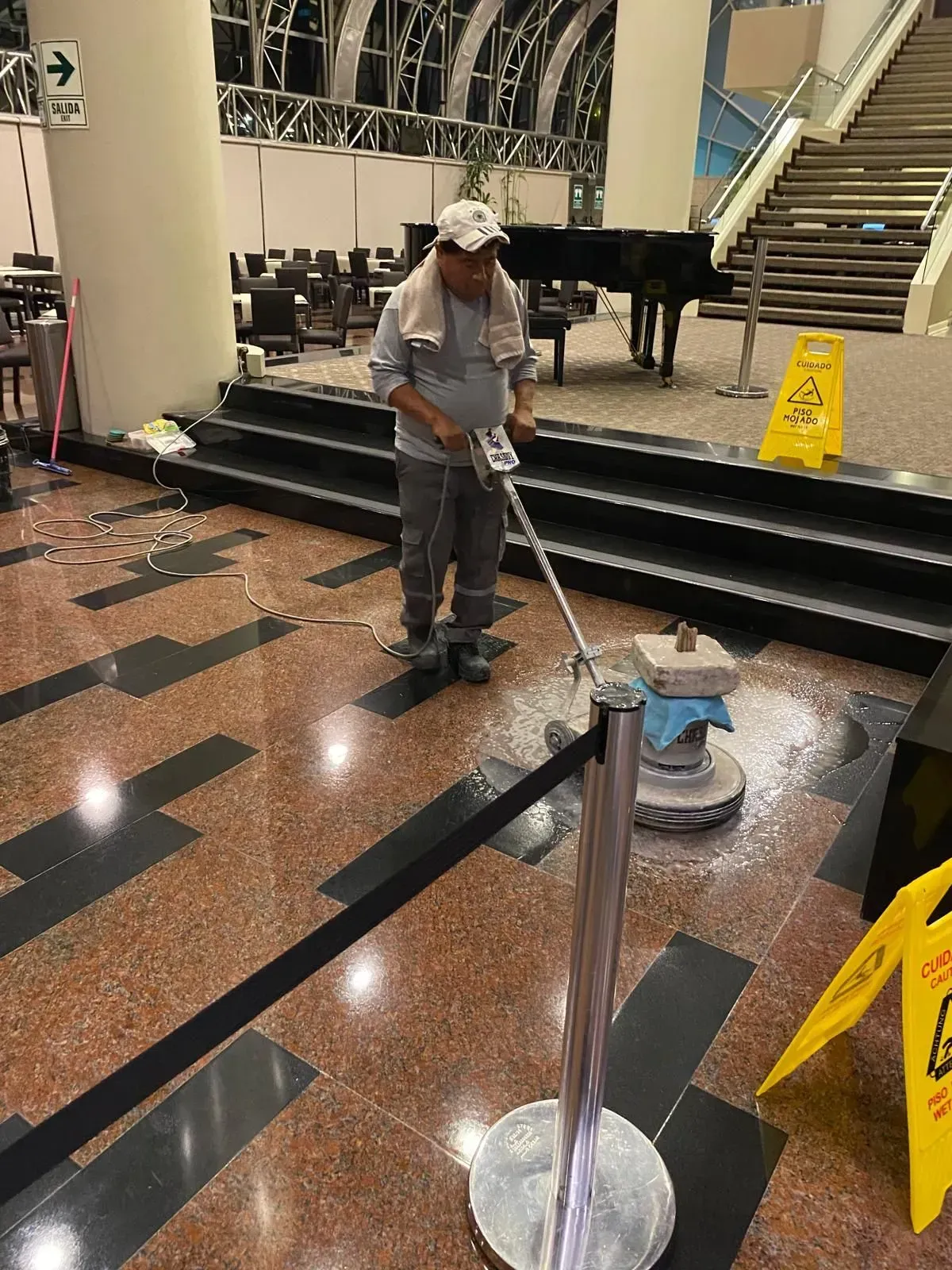 A person cleaning a red and black tile floor with a machine near a piano and caution signs.