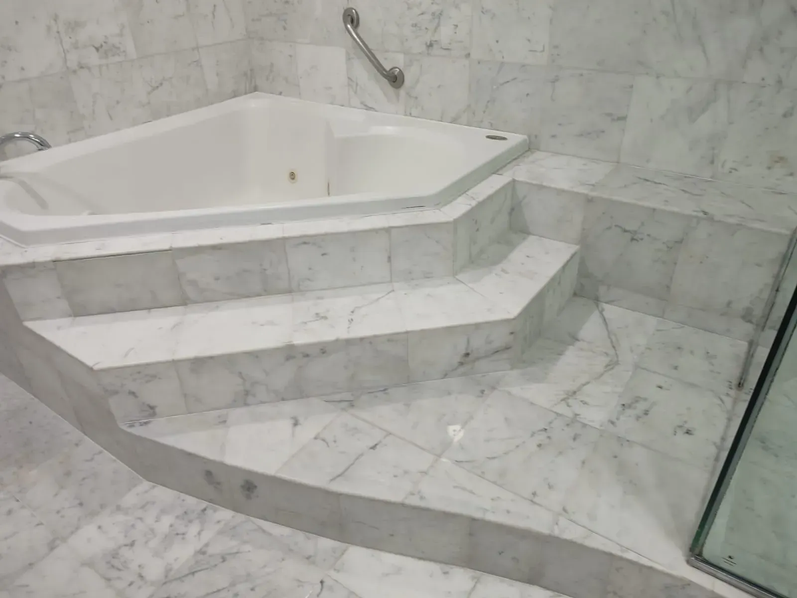White marble bathroom with triangular jacuzzi tub on raised platform with three steps.