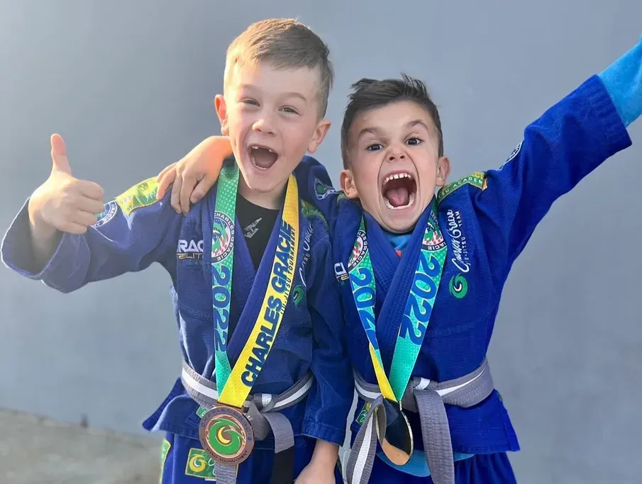 Two boys in blue martial arts uniforms, arms around each other, showing medals and cheering.