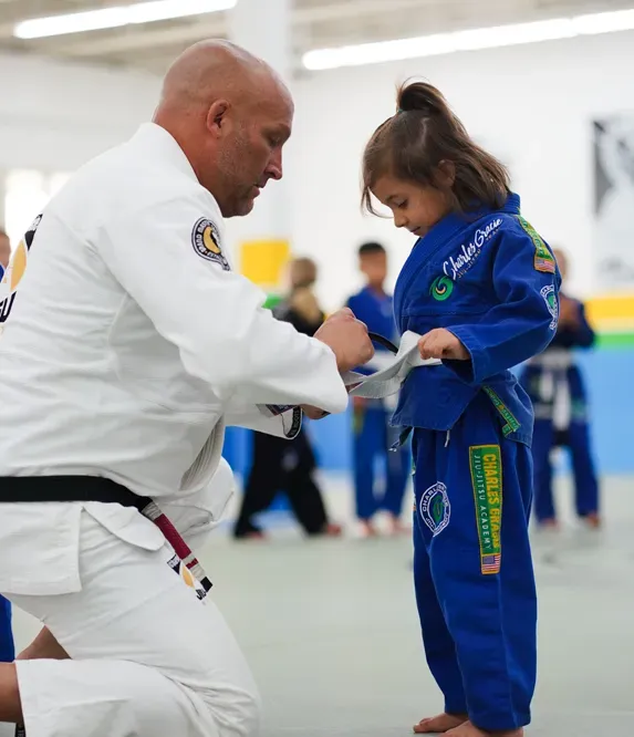 Man in white gi adjusts belt on a young child in a blue gi; they are in a gym.