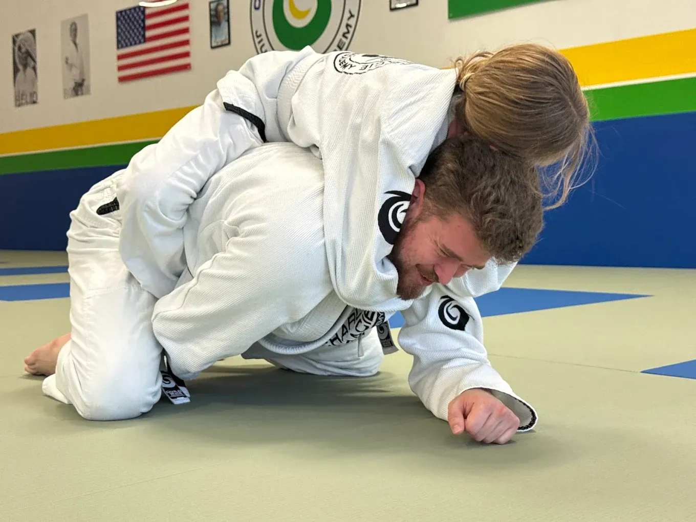 Person in white gi in a rear naked choke on another person in a white gi on a mat in a gym.