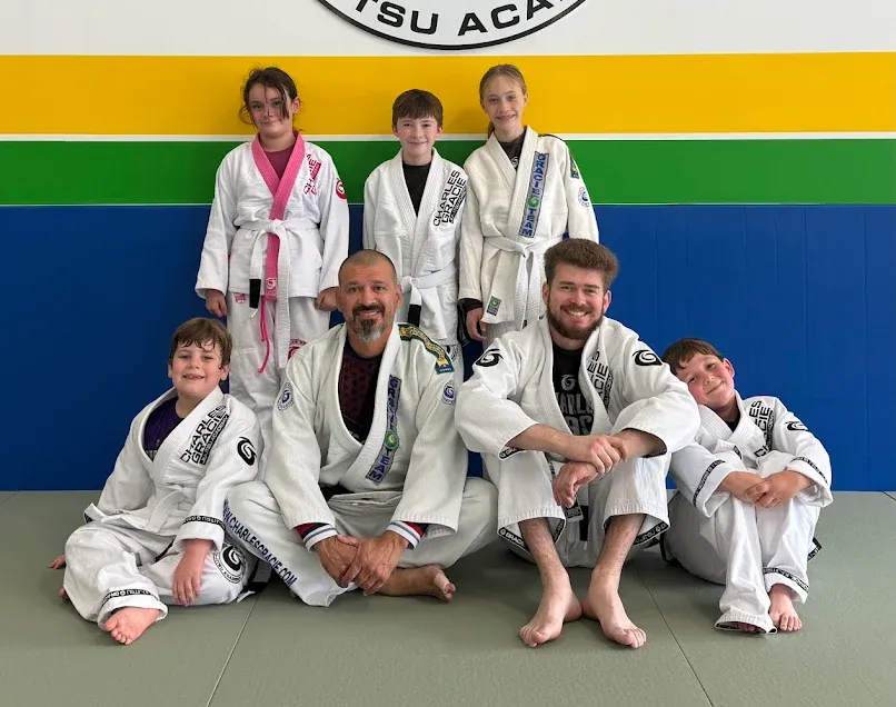 Group of people in martial arts uniforms, some with belts. Posing on a mat, near a blue, yellow and green wall.