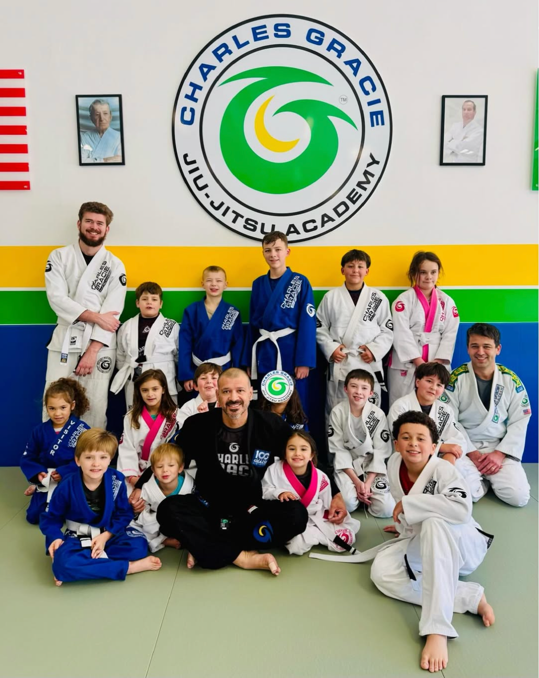 Group of children and adults in Jiu-Jitsu uniforms posing in front of a Charles Gracie Academy sign.