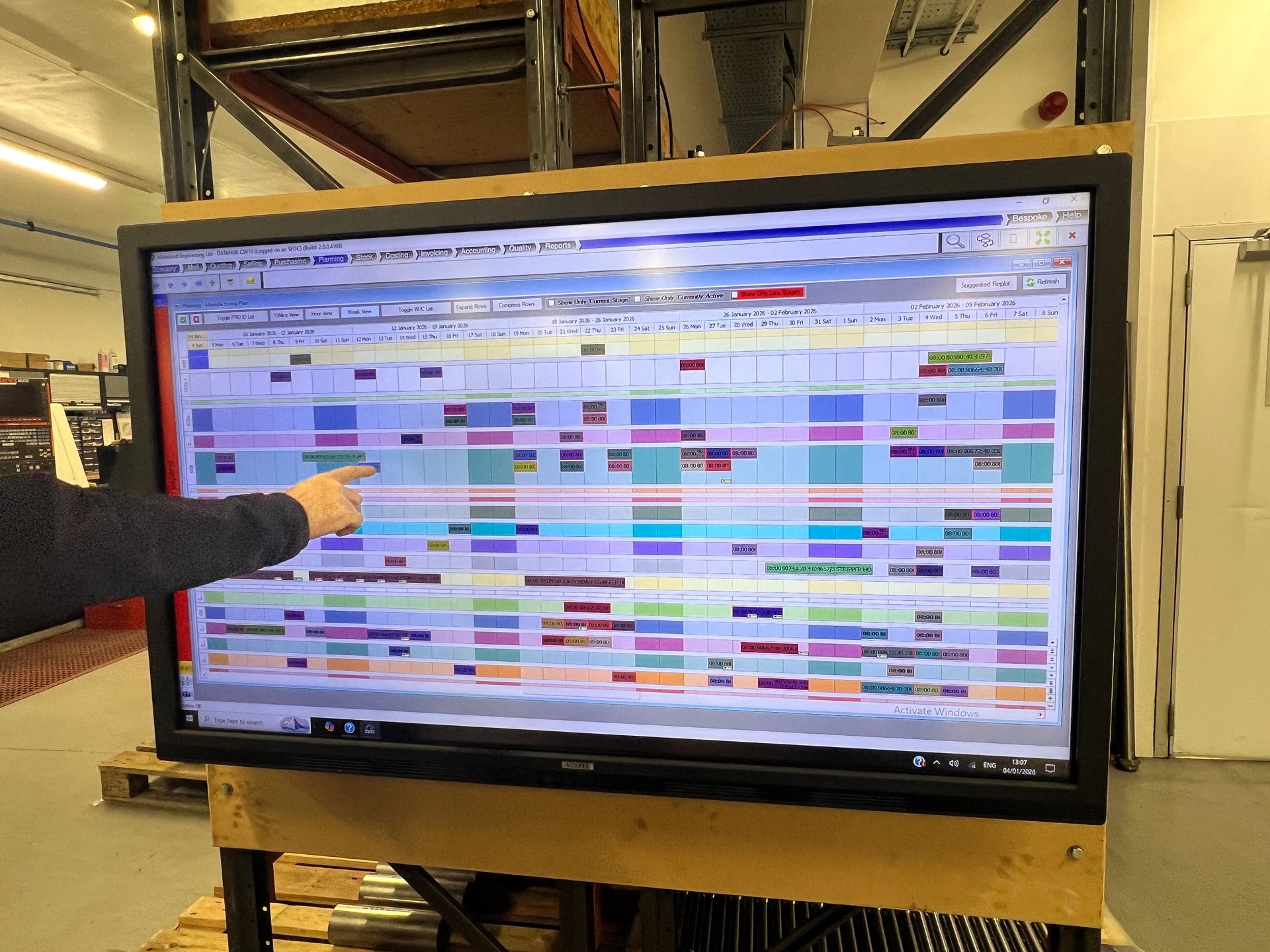 A hand points to a large digital display showing a complex project schedule with color-coded tasks in a warehouse setting.