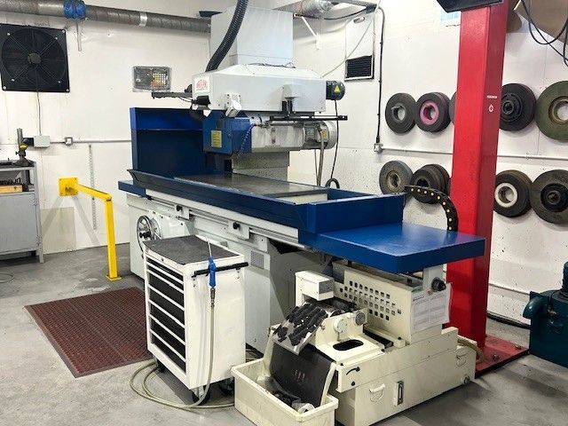 A blue and white industrial surface grinder stands in a machine shop with grinding wheels hanging on the wall.