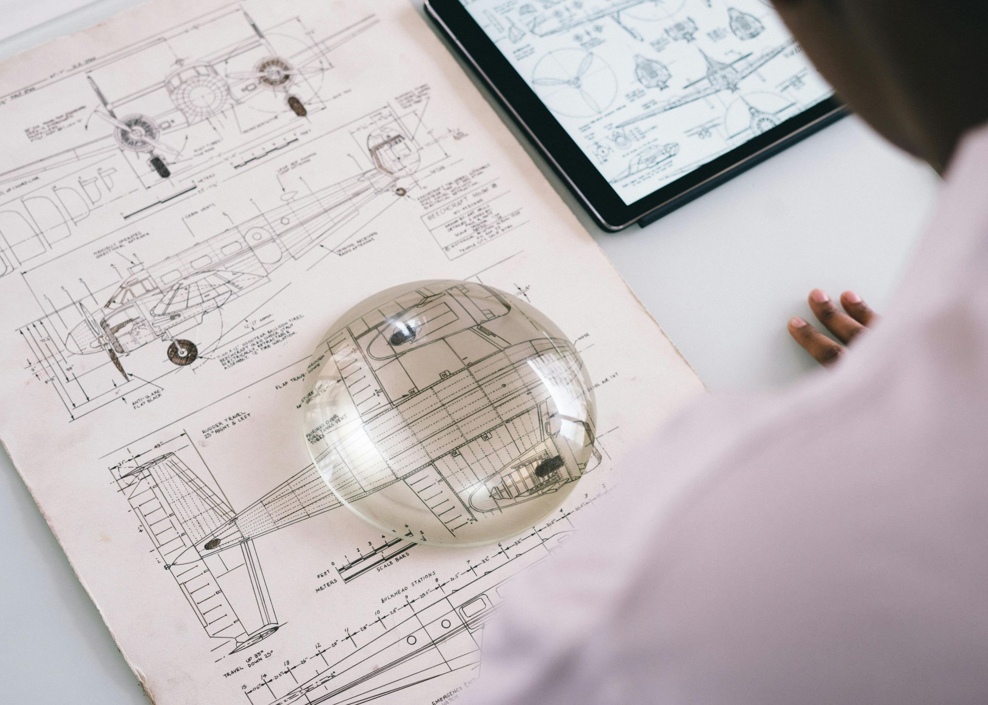 A person looks at engineering diagrams on paper with a dome magnifier, with a tablet showing similar schematics nearby.