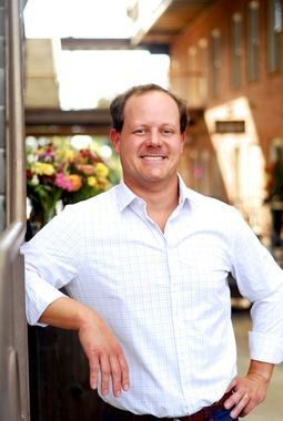 Man in a button-down shirt smiles, leaning against a wooden post. Floral arrangement in background.