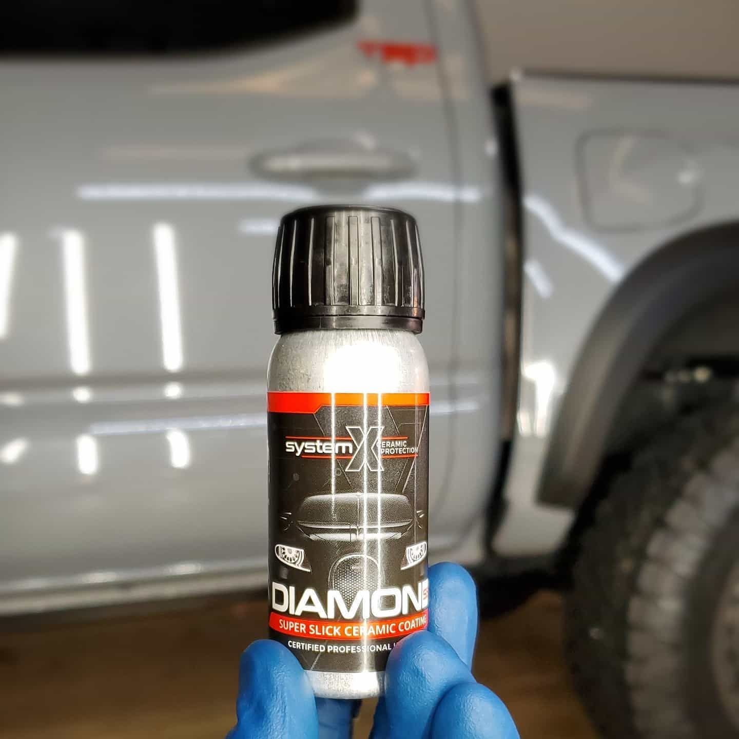 A person in blue gloves holds a System X Diamond ceramic coating bottle in front of a gray truck.