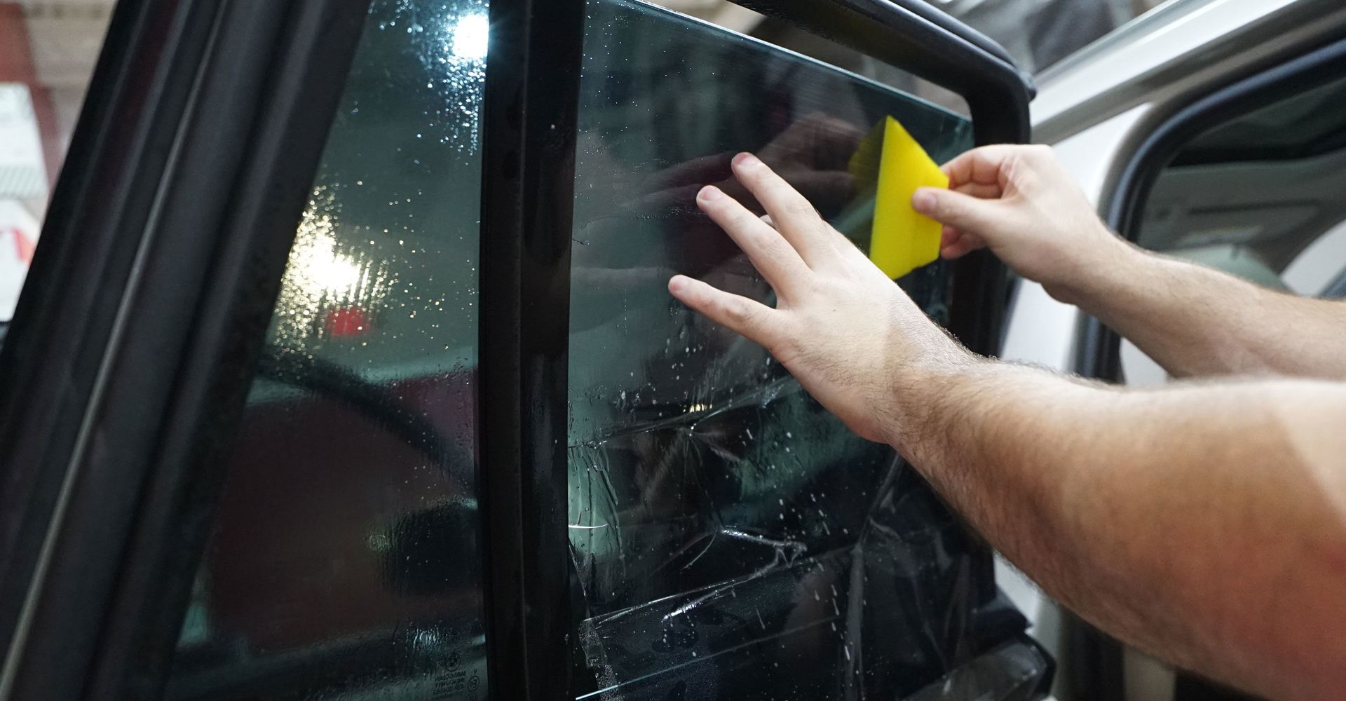 Person applying window tint to a car window with a yellow squeegee.