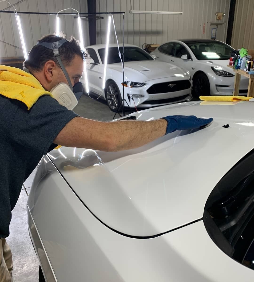 Man in mask and gloves detailing a white car. Another white car and a Tesla are in the background.