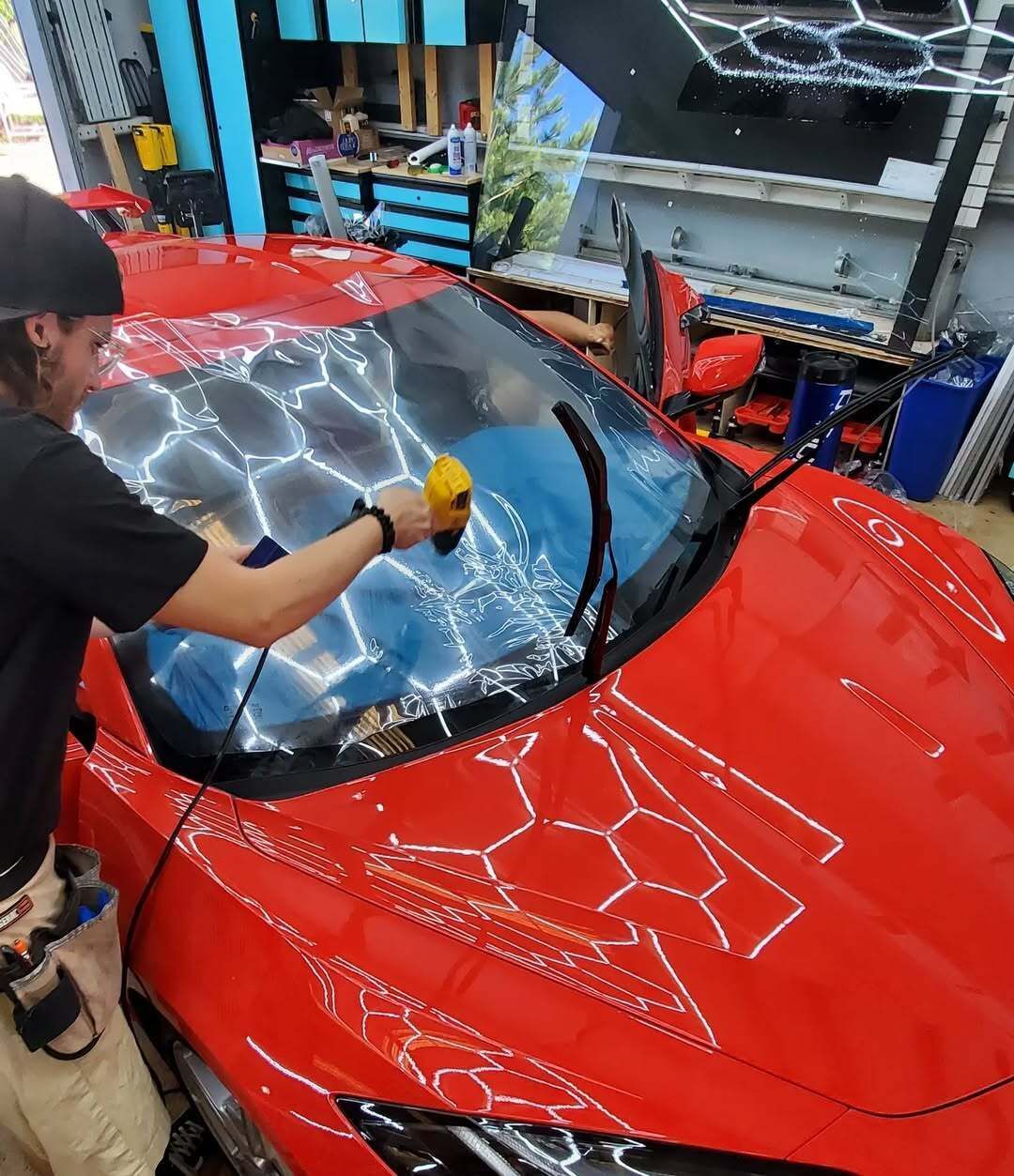 Person applying film to a red car windshield with a heat gun. Workshop setting.