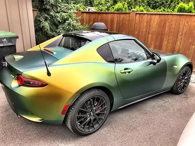 Green and gold color-shifting Mazda Miata sports car parked on a paved surface next to a wooden fence.