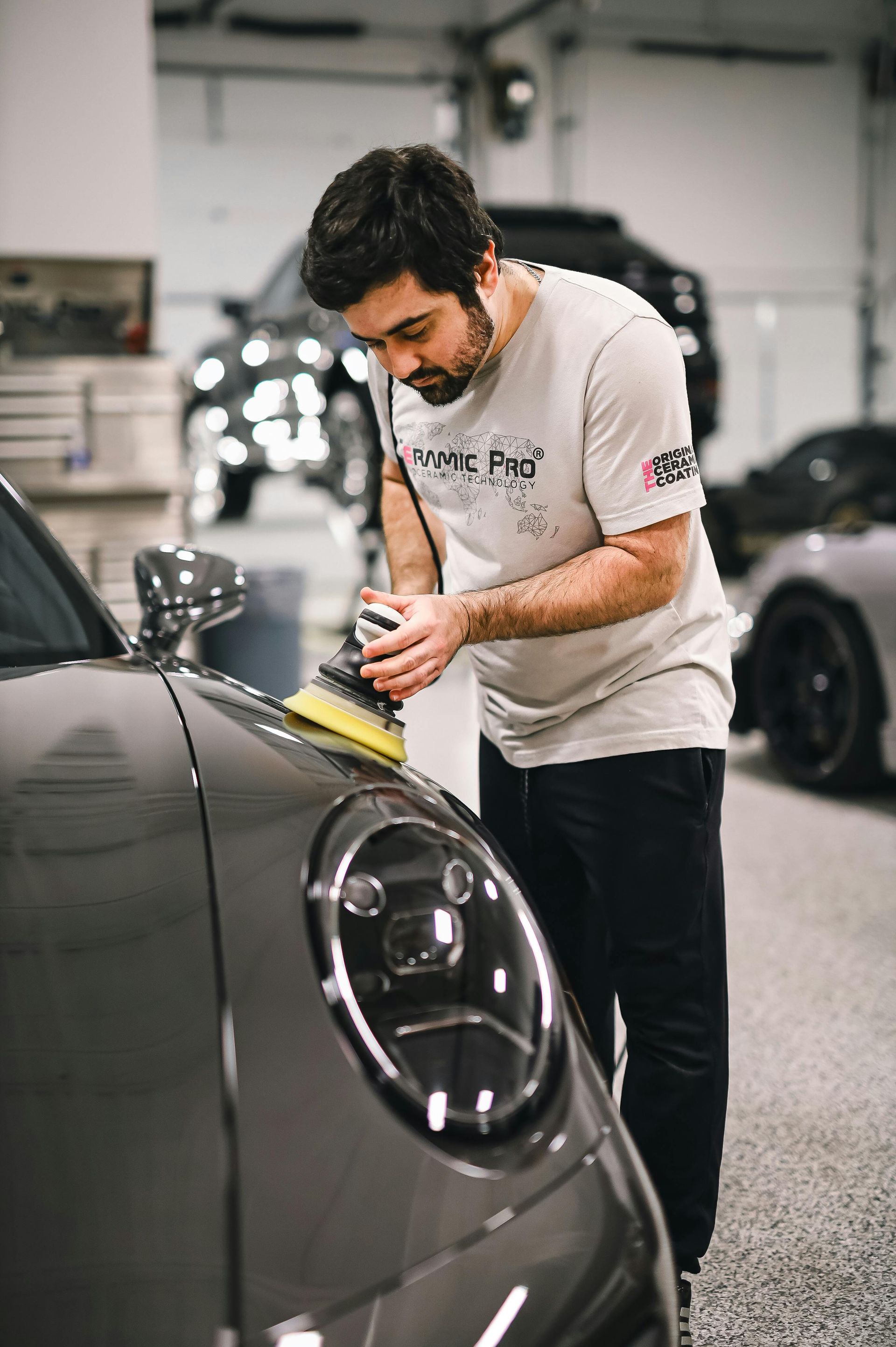 A person in a gray t-shirt uses a yellow power polisher on the hood of a dark gray luxury car in a professional garage.