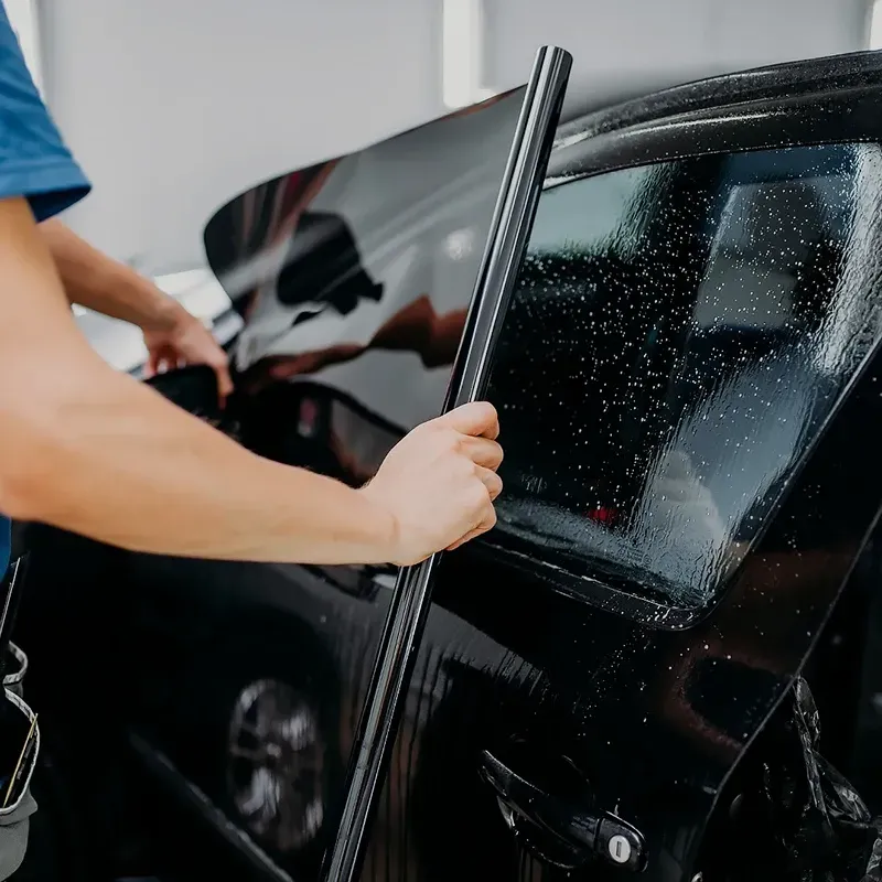 Person applying tint film to a black car window in a garage.