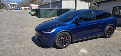 A blue Tesla Model X parked on asphalt, with black wheels and red brake calipers, under a sunny sky.