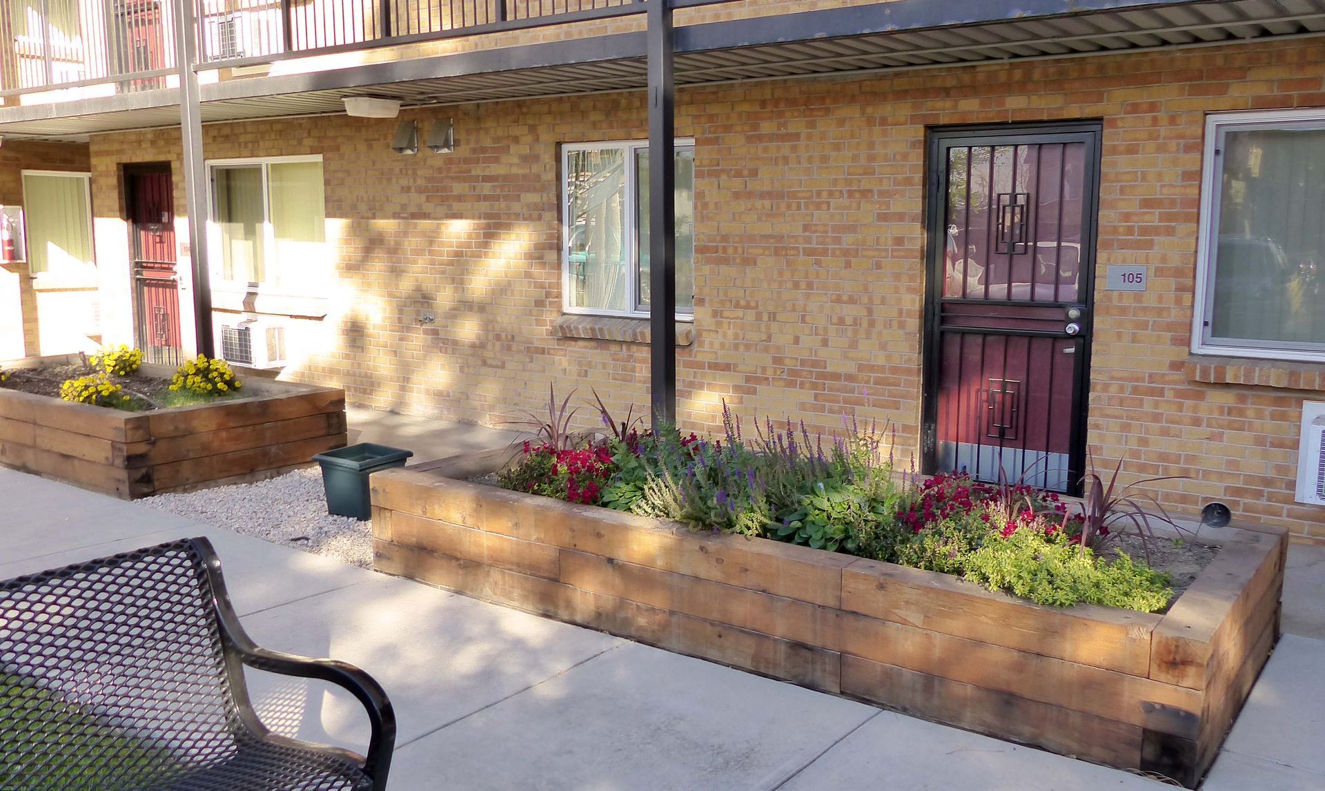 A yellow brick apartment building exterior features wooden raised flower beds, a black metal bench, and gated doorways.