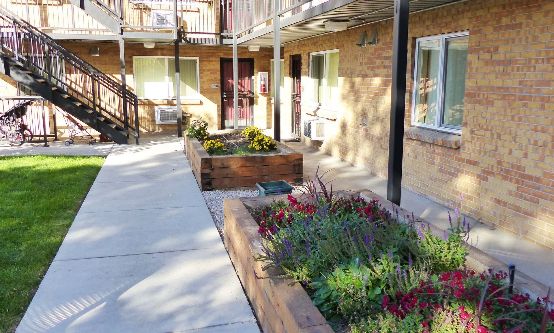 A concrete walkway leads past two raised wooden planter beds filled with colorful flowers near a brick apartment building.