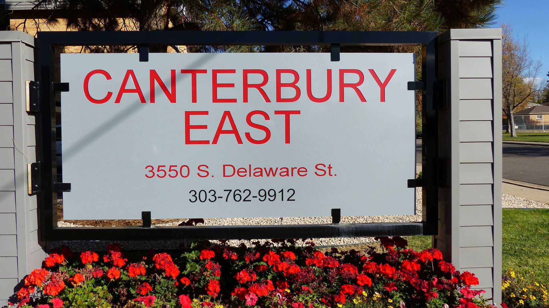 A sign for Canterbury East apartments at 3550 S. Delaware St., phone 303-762-9912, behind a bed of red flowers.
