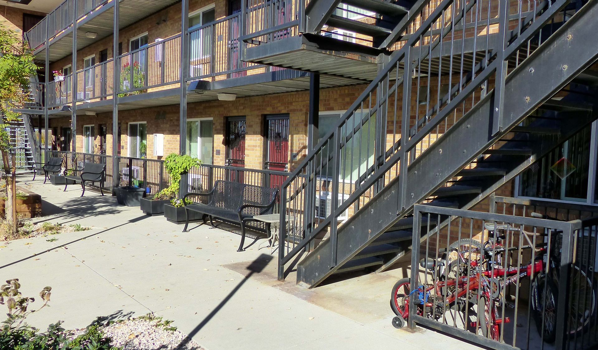 A brick apartment building exterior with a metal staircase, walkway, benches, and a bike rack on a sunny day.