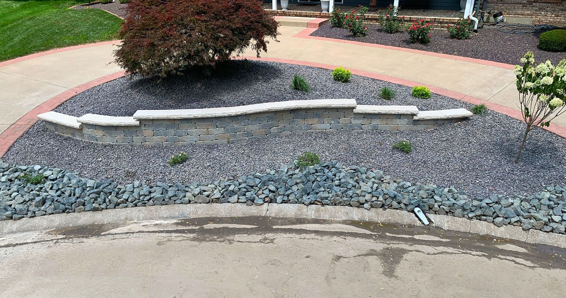 A decorative landscaping island with dark gravel, a low stone wall, a red bush, and small plants on a concrete driveway.