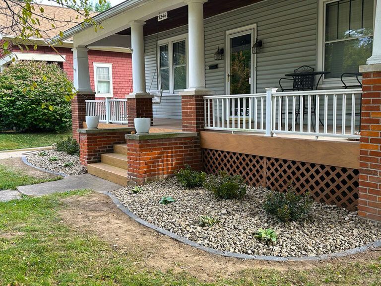 A house with a large front porch featuring white pillars, red brick details, white railings, and a stone garden bed.
