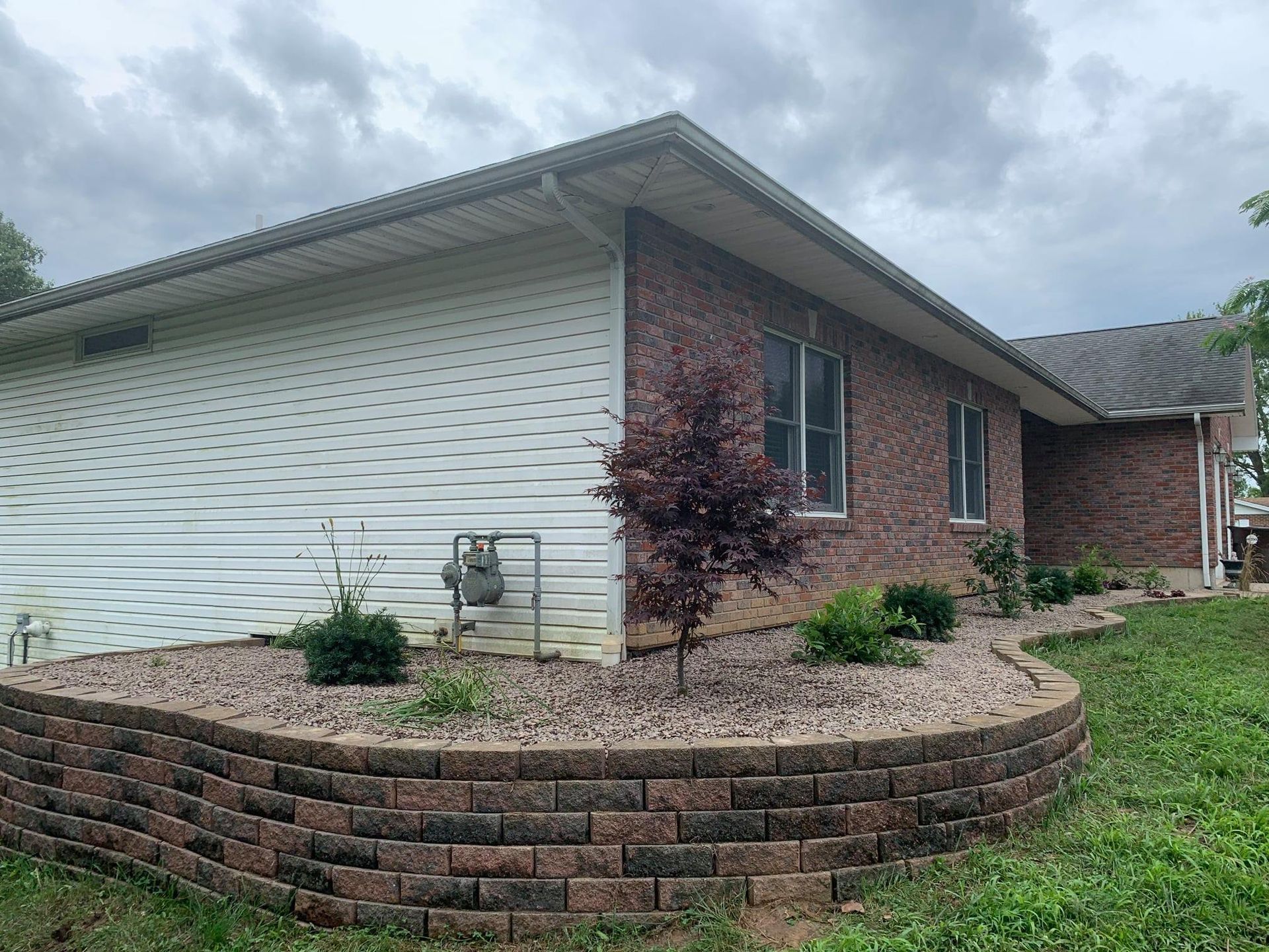 A brick and white-sided house with a curved, tiered stone retaining wall filled with gravel and small shrubs.