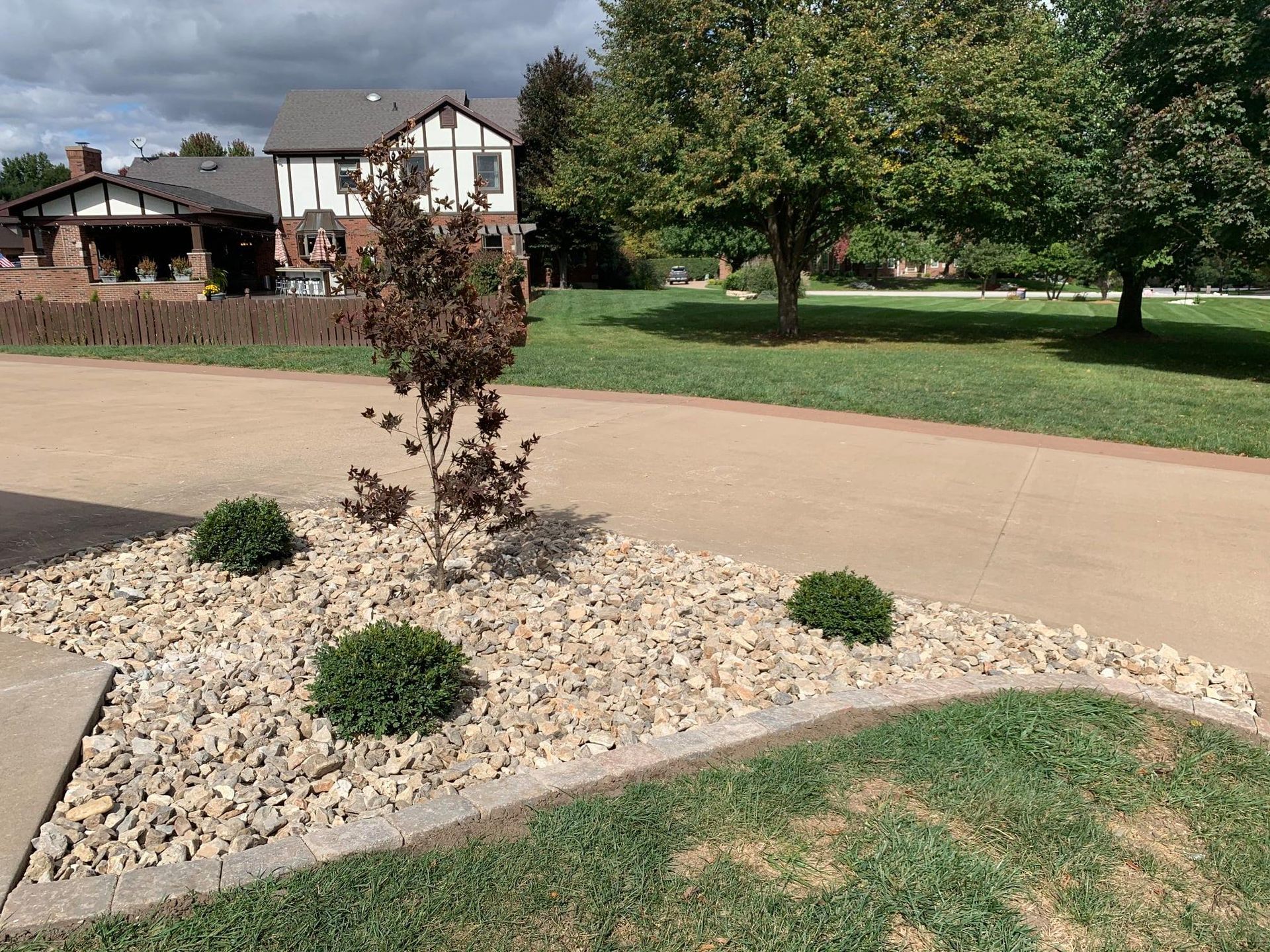 A landscaped garden bed with small green shrubs and a purple-leafed tree, surrounded by light-colored rocks near a driveway.