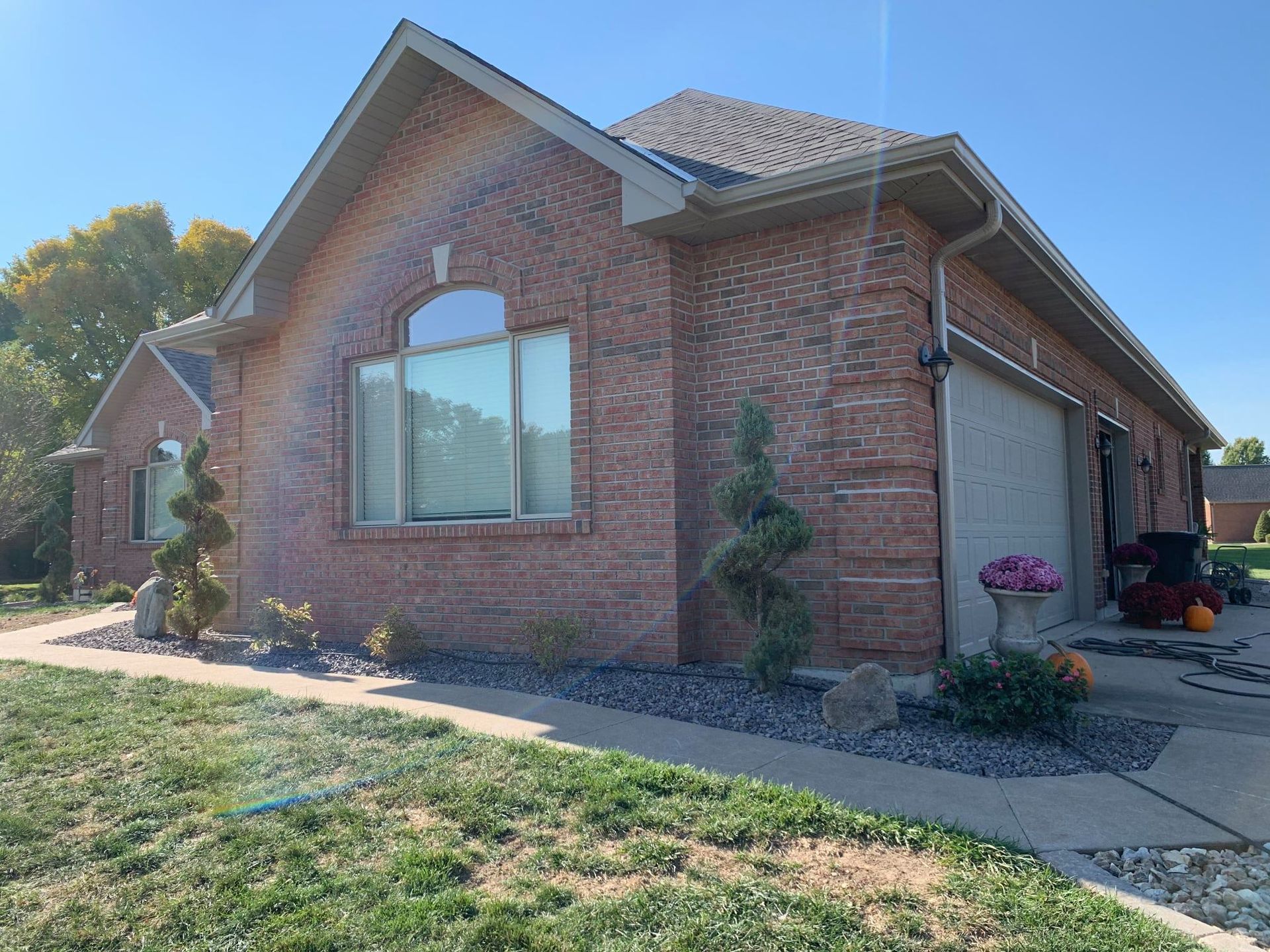 A brick ranch house with a large window and attached garage, featuring spiral shrubs in a garden bed on a sunny day.