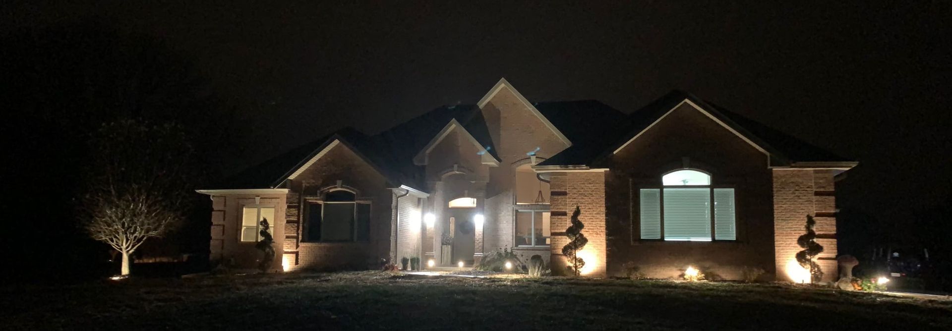 A brick house at night, illuminated by warm outdoor landscape lighting against a dark sky.