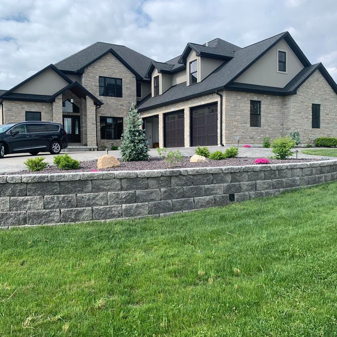 A large, two-story stone house with a dark roof and three garage bays, viewed from a green lawn with a retaining wall.