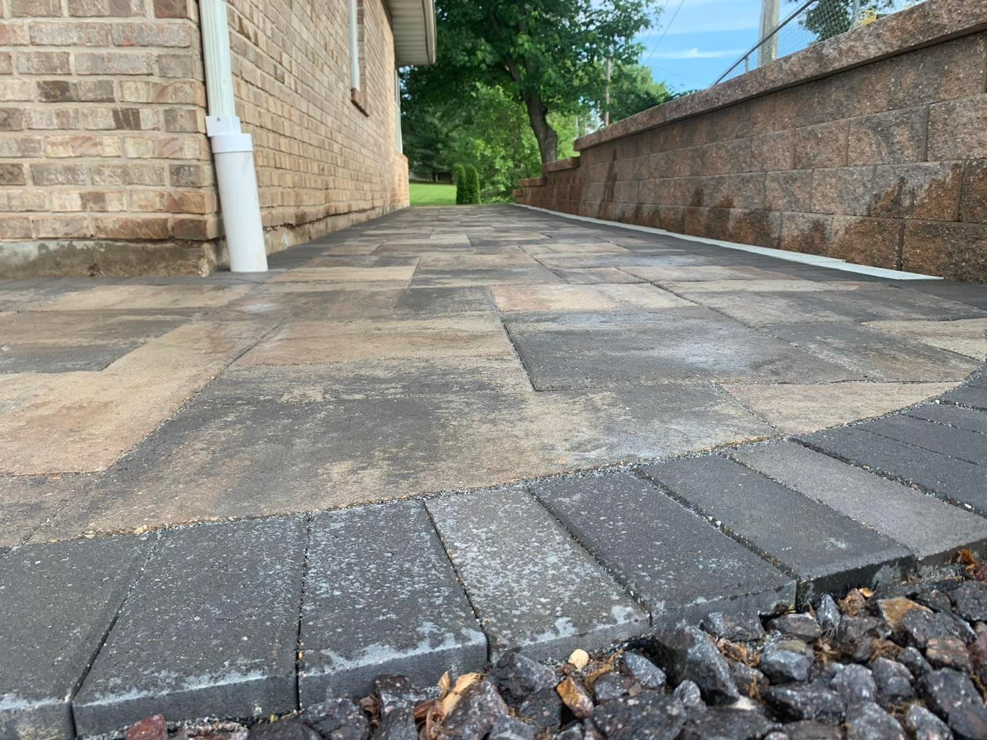 A ground-level view of a paver walkway with a dark brick border, situated between a brick house wall and a stone wall.