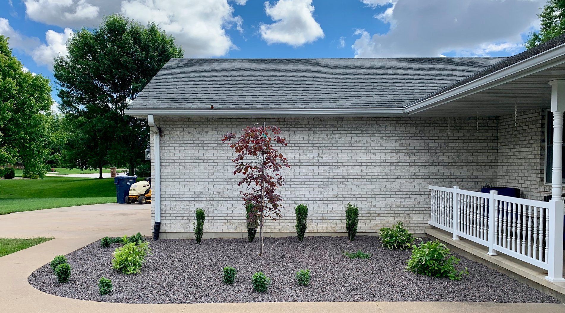 A house exterior with white brick, a gray roof, a porch railing, and a front landscaping bed with dark stone and plants.