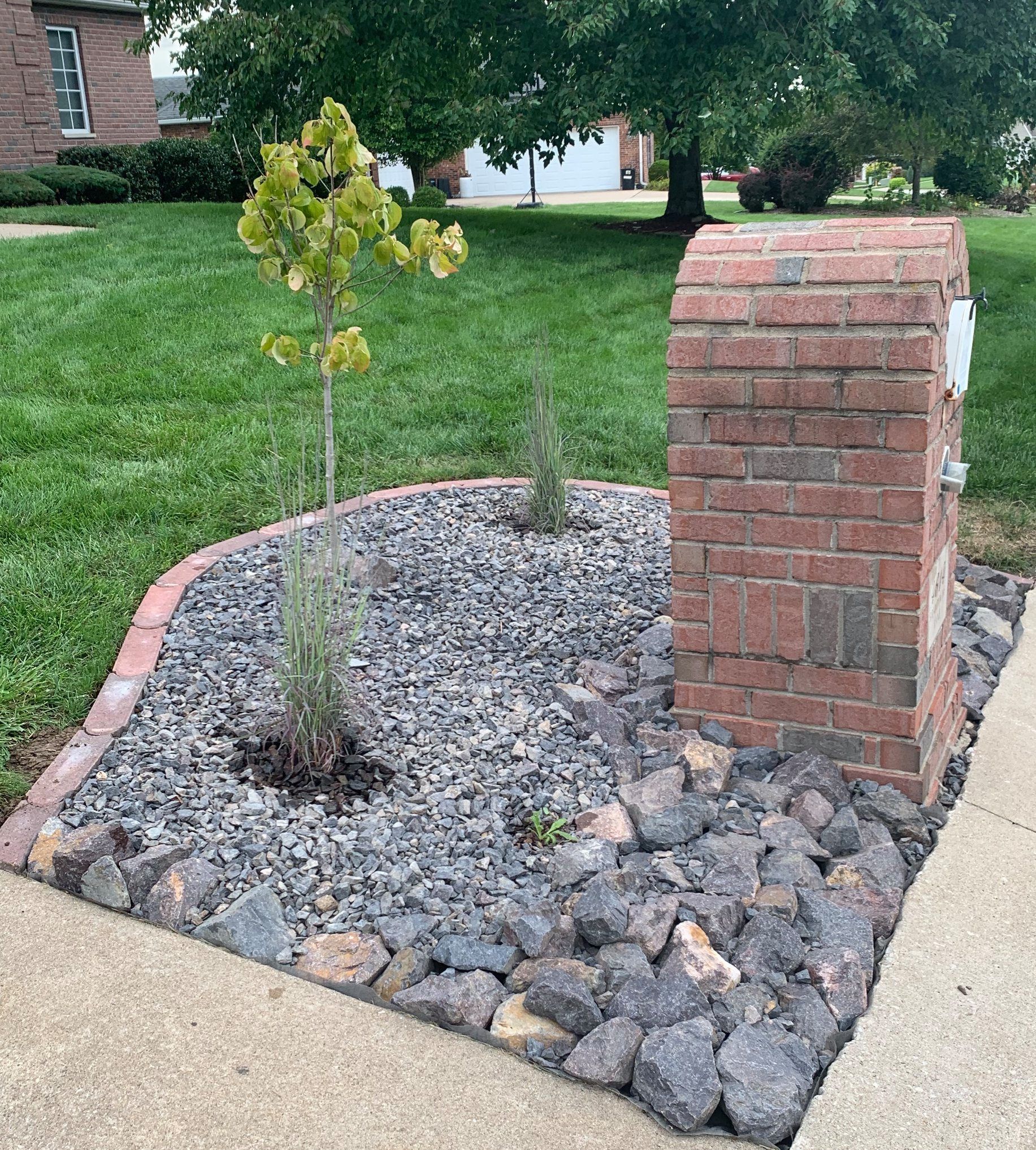 A brick mailbox sits in a gravel garden bed with a small green tree, bordered by bricks, next to a concrete sidewalk.