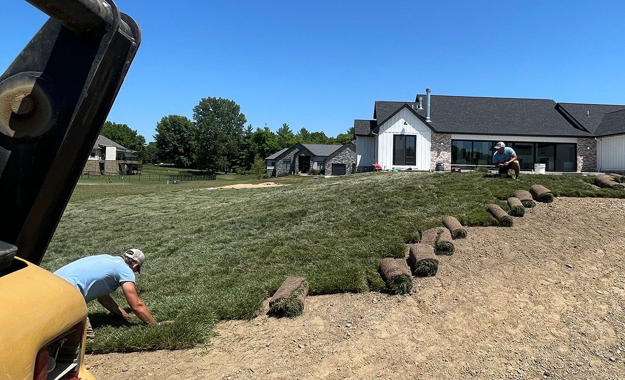 Two workers lay sod on a sloped residential lawn on a sunny day, near a large house with a black roof.