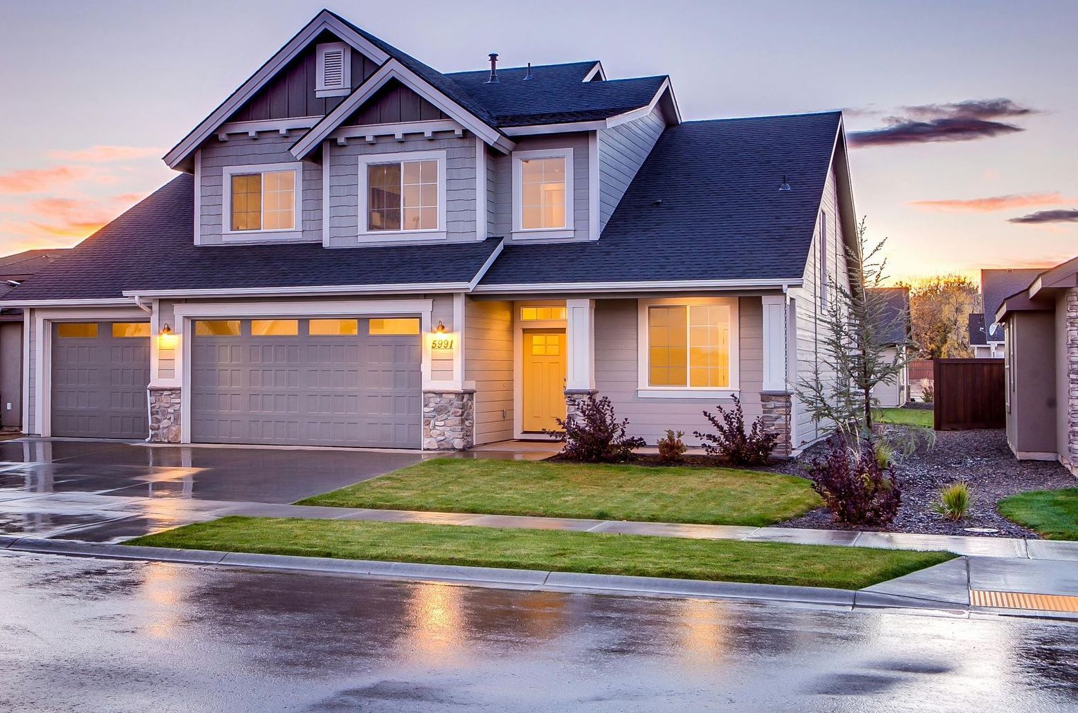 Two-story gray suburban house with lit windows and a wet driveway at sunset