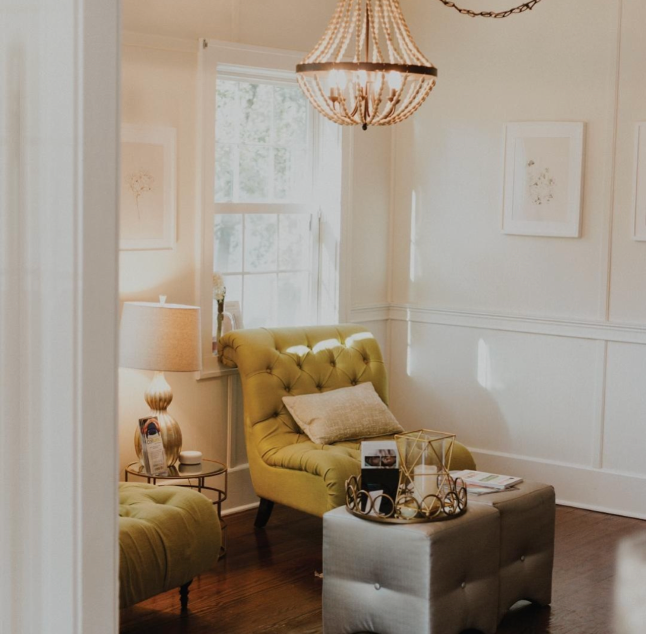 Cozy boutique day spa client waiting area with green chairs and and ottoman, gold accents, chandelier, and framed art.