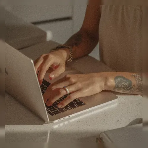 Person typing on a laptop with a tattooed arm, sunlight on white surface.