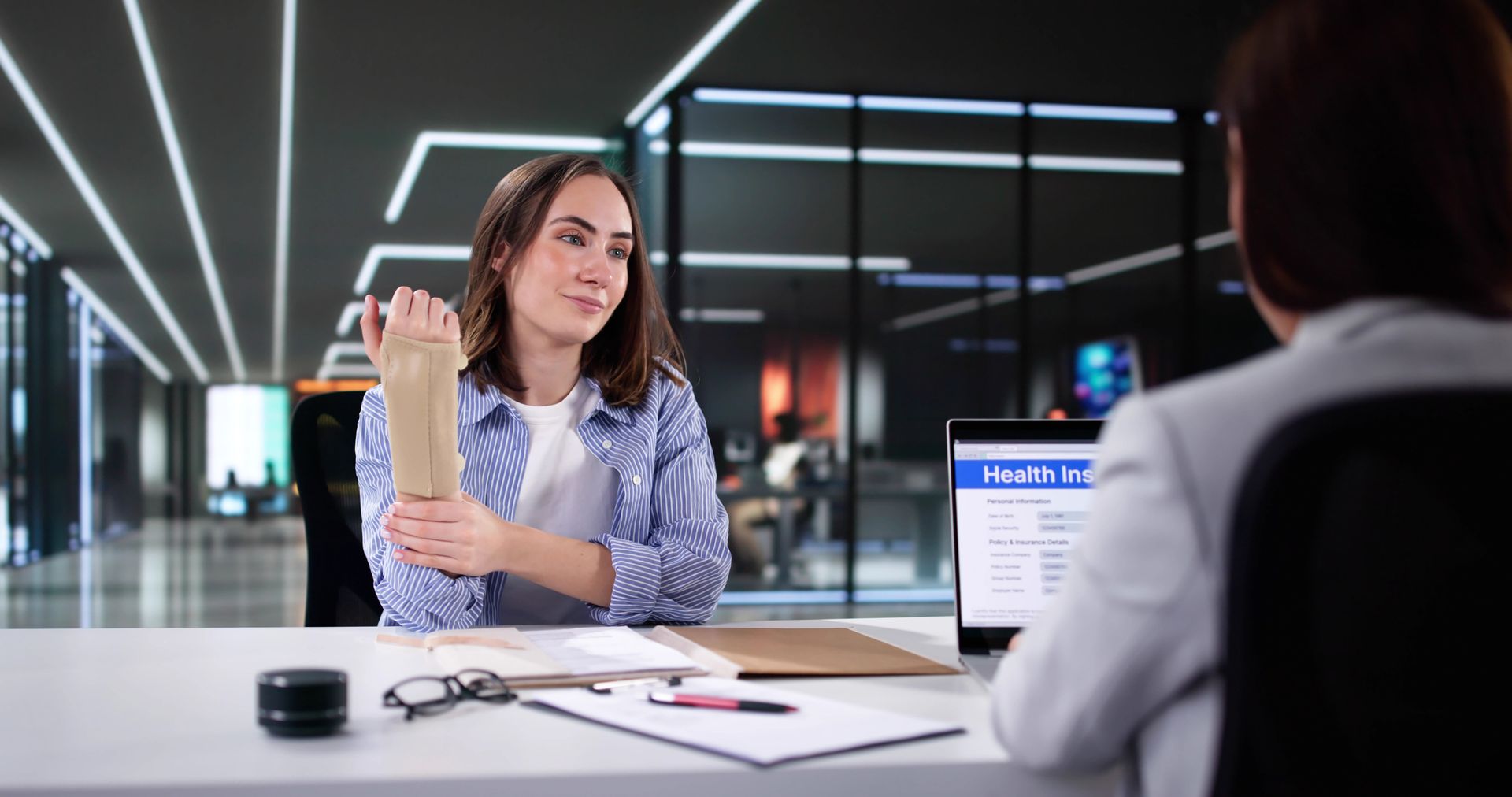 Woman gesturing while talking, sitting across from another person at a desk in an office. Laptop screen visible.