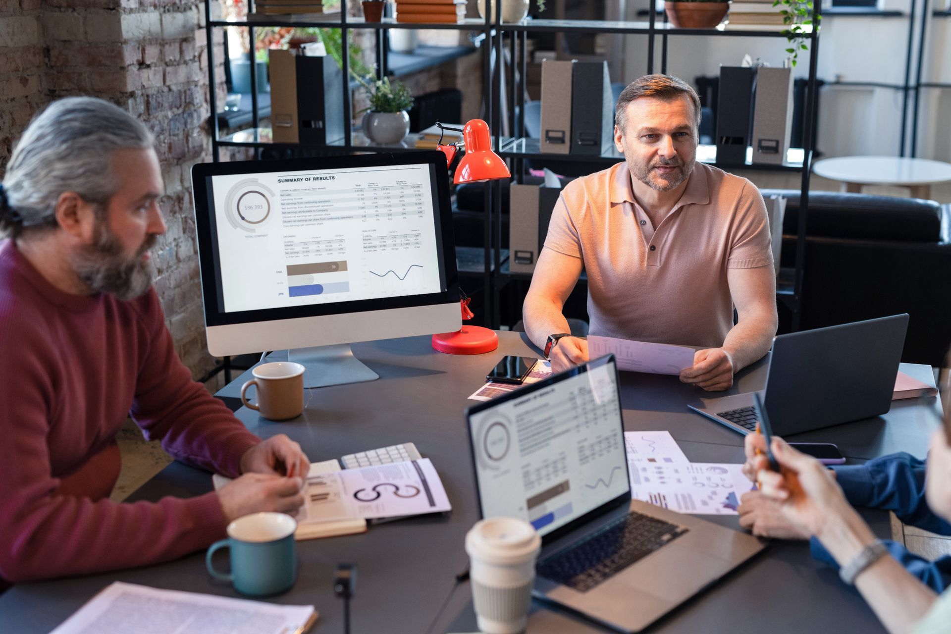 Four people in a meeting around a table with laptops, papers, and coffee in a modern office.