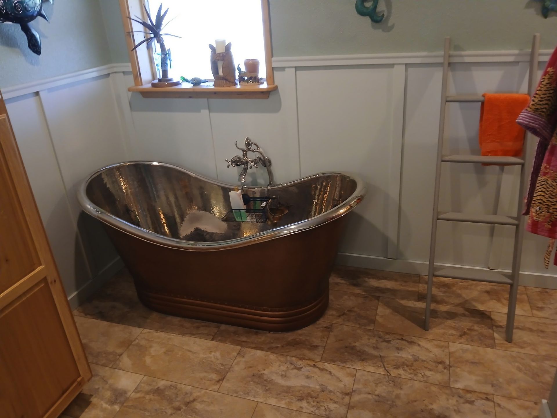 Copper bathtub in a bathroom with a window, ladder, and wood paneling.